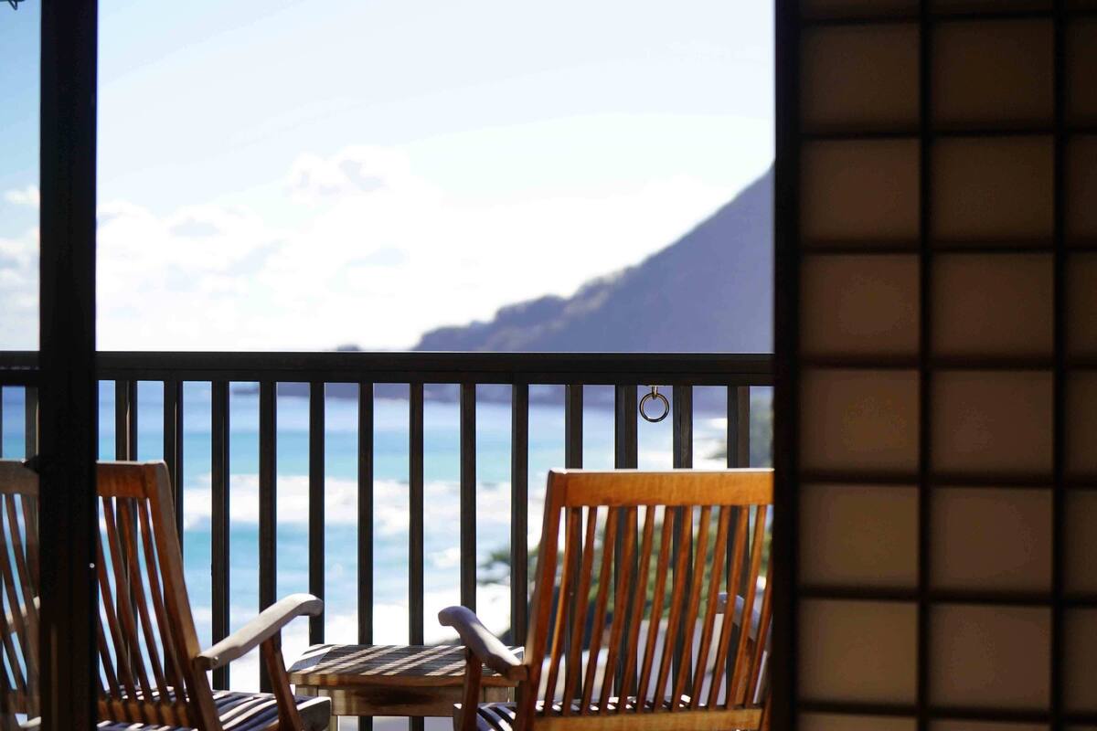Un balcón con dos sillas de madera y una pequeña mesa, con vista a una playa y una montaña al fondo.