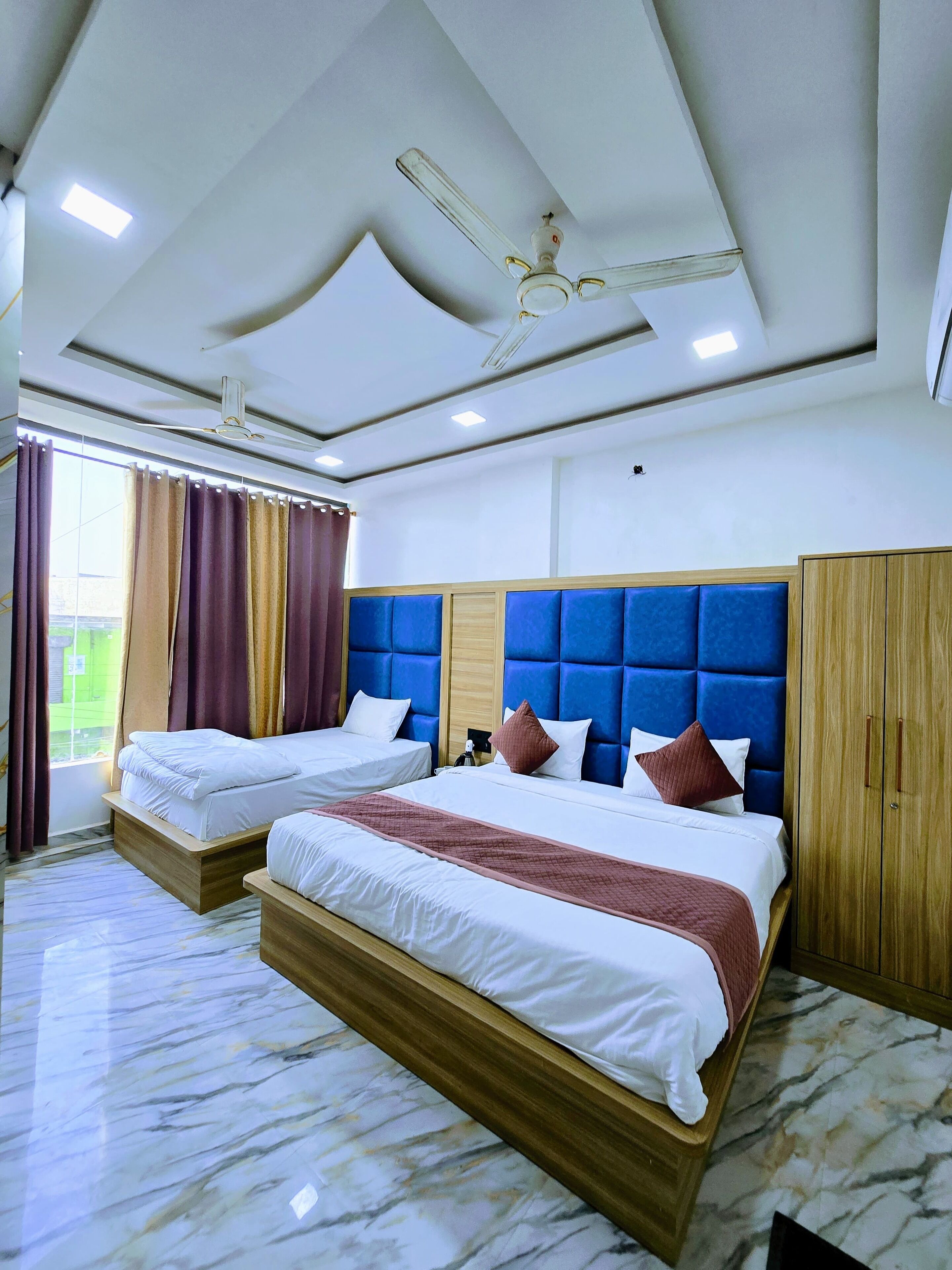 A hotel room with two beds, a blue headboard, wooden wardrobe, and a ceiling fan.