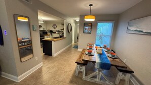 A modern kitchen with a central island, a dining area with a long table set for six, and a view of the outdoors through a window.