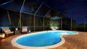Ein Schwimmbad mit Glasüberdachung, umgeben von Pflasterböden und Korbmöbeln mit Kissen.