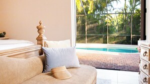 Ein Schlafzimmer mit Bett, Sofa und Kommode. Ein Fenster bietet Blick auf einen Swimmingpool und die umliegende Natur.
