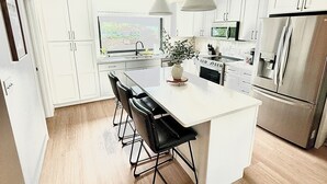 A modern kitchen with white cabinetry, a central island with bar stools, stainless steel appliances, and pendant lighting. - Modern 3-Story Condo Across from Arnold’s Park – Walk to Fun, Lake, & Dining! (Arnolds Park)