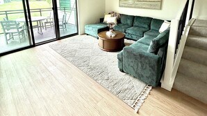 A spacious living room with a large sliding glass door leading to a deck, a green sofa, a round coffee table, and a ceiling fan. - Modern 3-Story Condo Across from Arnold’s Park – Walk to Fun, Lake, & Dining! (Arnolds Park)