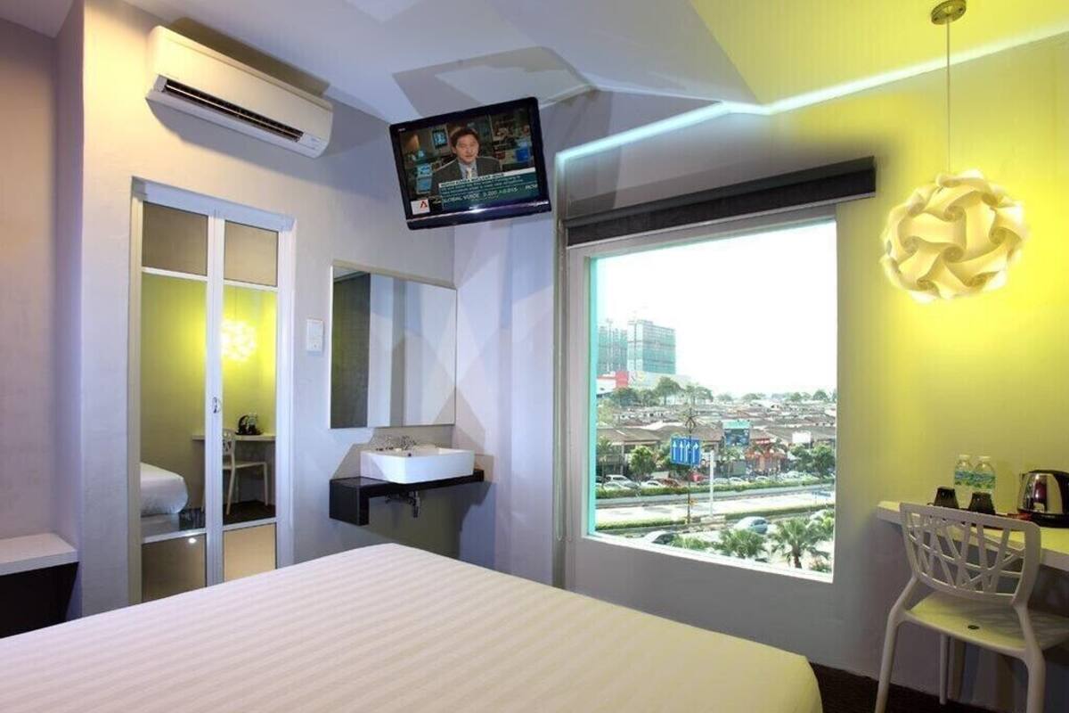 Habitación de hotel con cama, escritorio, silla, televisor, ventana con vista a la ciudad y baño con espejo y lavamanos.