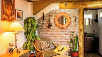 Una habitación acogedora con pared de ladrillo, techo de madera, espejo redondo, sillón mecedor, lámpara y plantas en macetas.