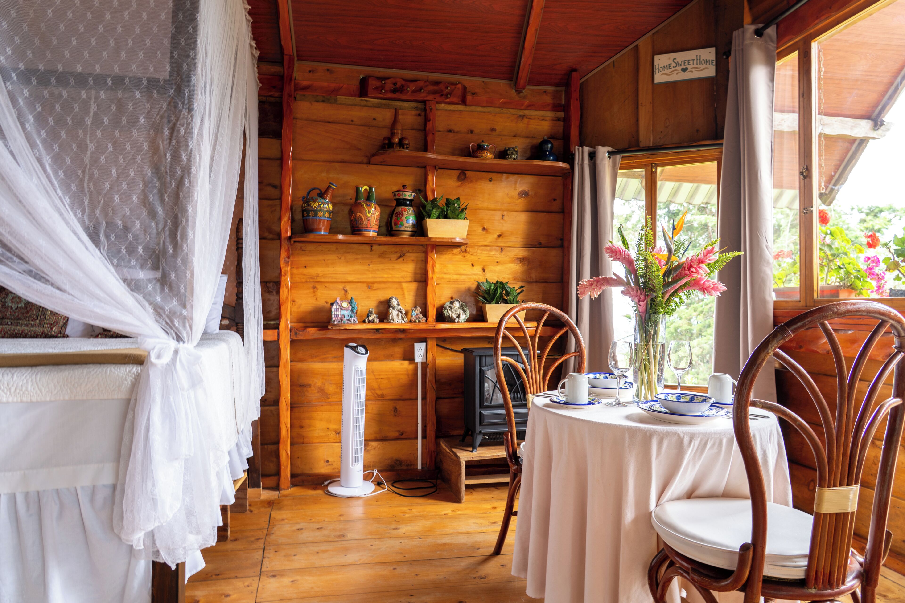 A wooden cabin interior with a dining table, chairs, a heater, and a window with a view of greenery.