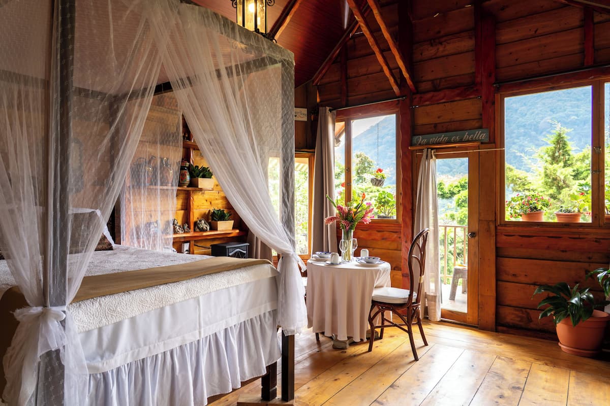 Une cabane en bois avec un lit, une petite table et des chaises, un balcon agrémenté de plantes en pot et une vue sur la verdure.