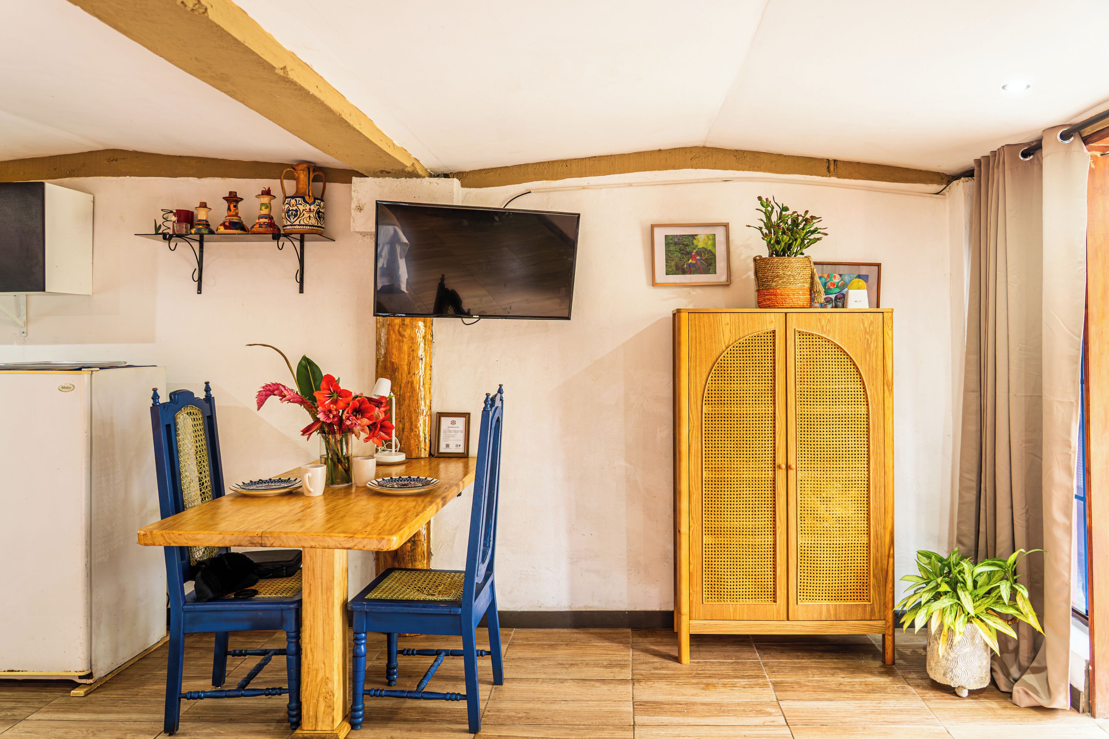 Un comedor con una mesa de madera, sillas azules, un televisor montado en la pared y un armario de mimbre.