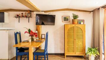 Un comedor con una mesa de madera, sillas azules, un televisor montado en la pared y un armario de mimbre.