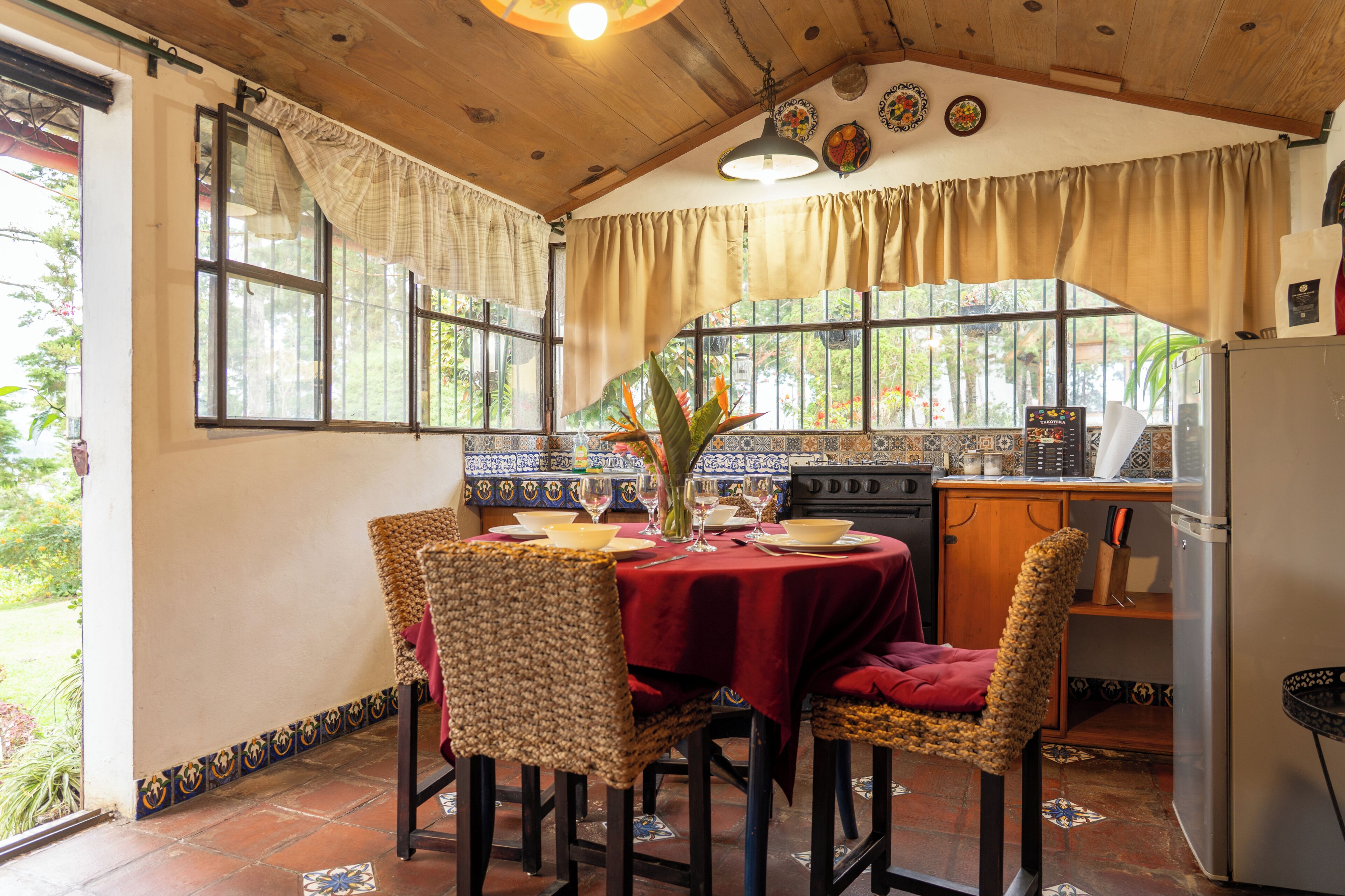 A dining area with a table set for four, surrounded by wicker chairs, a red tablecloth, and a kitchen in the background.