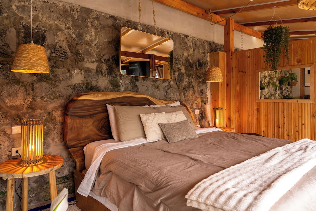 Une chambre à coucher au style rustique, avec un mur en pierre, des poutres en bois, un lit agrémenté de nombreux coussins et des lampes suspendues.