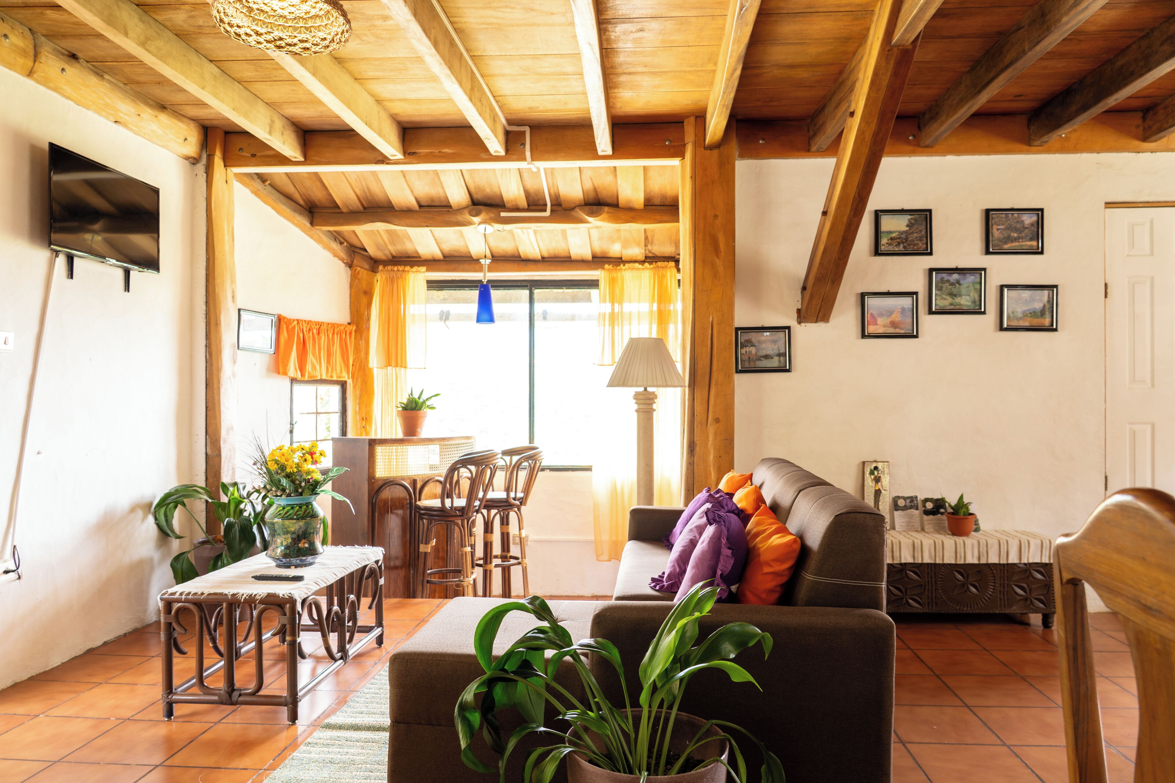 A living room with a wooden ceiling, a sofa, a table with chairs, and a TV mounted on the wall.