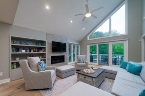 A spacious living room with a fireplace, comfortable seating, a flat-screen TV, and large windows offering a view of the outdoors.