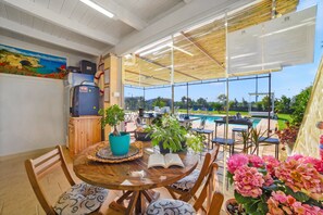 A sunlit patio with a pool, wooden furniture, and potted plants.