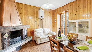 Una habitación acogedora con chimenea, paredes de madera, un sofá blanco, una mesa de comedor con un cuenco de fruta y una ventana con vistas.