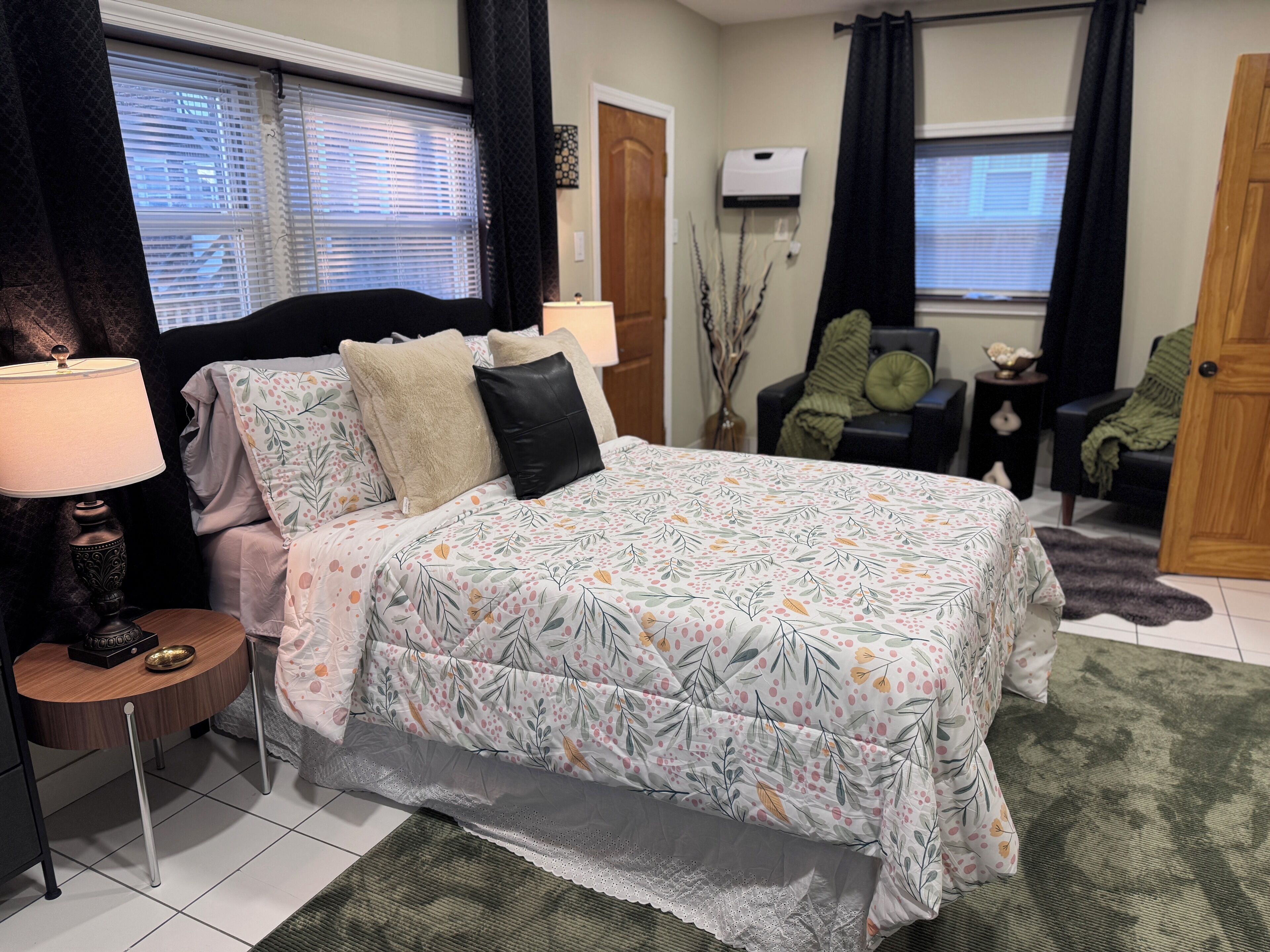 A bedroom with a bed, bedside tables, a lamp, a chair, a door, and a window with blinds.