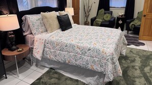 Ein Schlafzimmer mit einem Bett, Nachttischen, einer Lampe, einem Sessel, einer Tür und einem Fenster mit Jalousien.