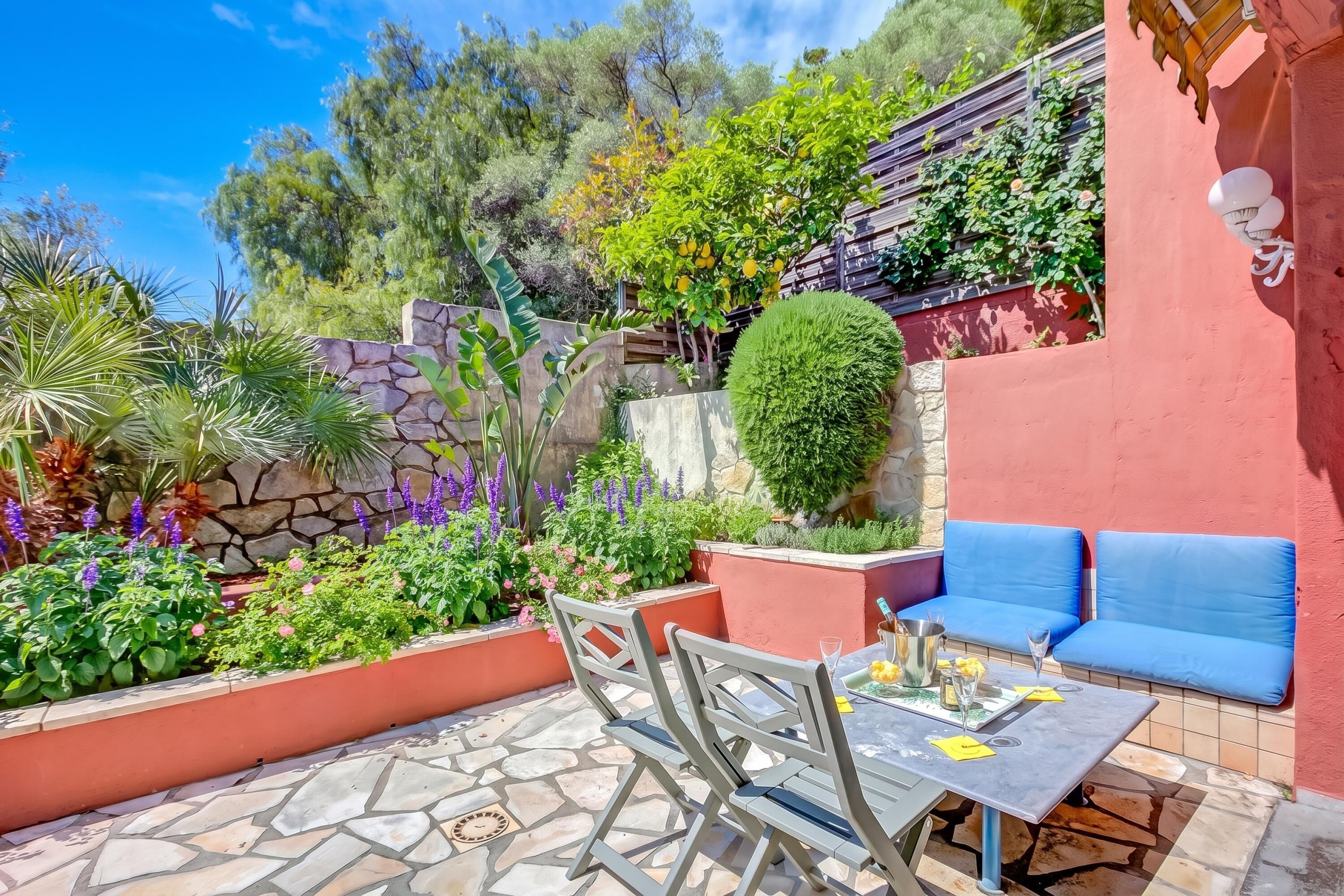 An outdoor seating area with stone flooring, a table with chairs, and a variety of plants and flowers.