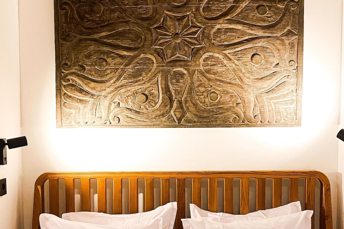 Un lit avec une literie blanche, une tête de lit en bois et une couvre-lit verte. Au-dessus du lit, on peut voir une œuvre d’art fixée au mur, dont le motif rappelle un arbre.