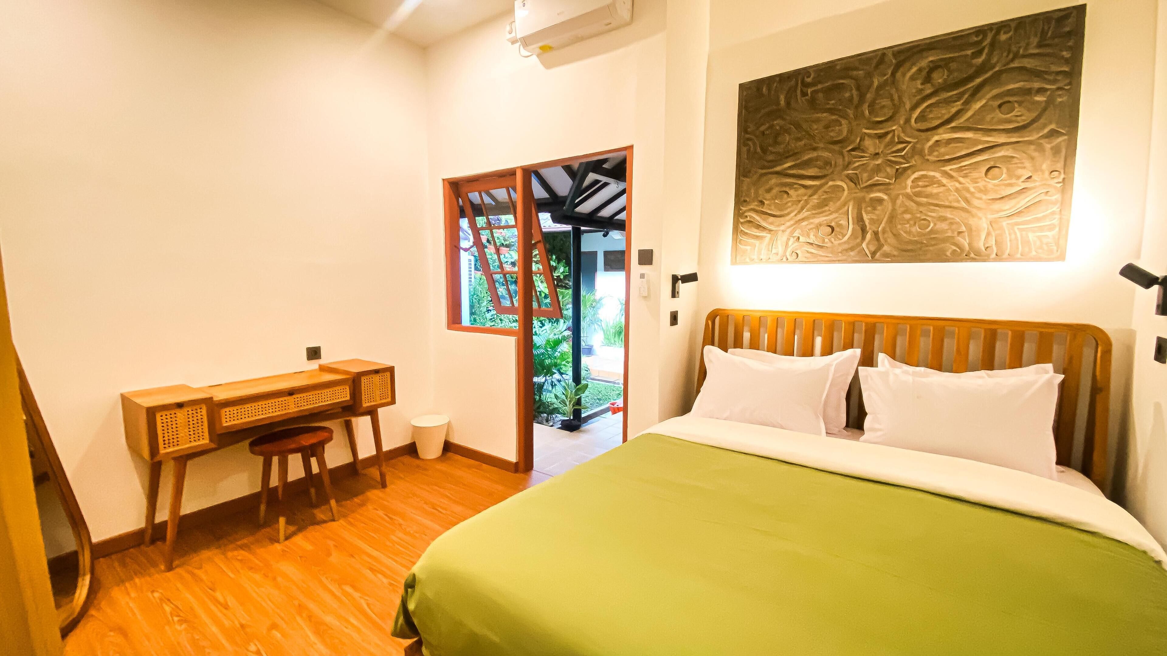 A bedroom with a bed, a desk, and a chair. There is a door leading to a balcony with plants.