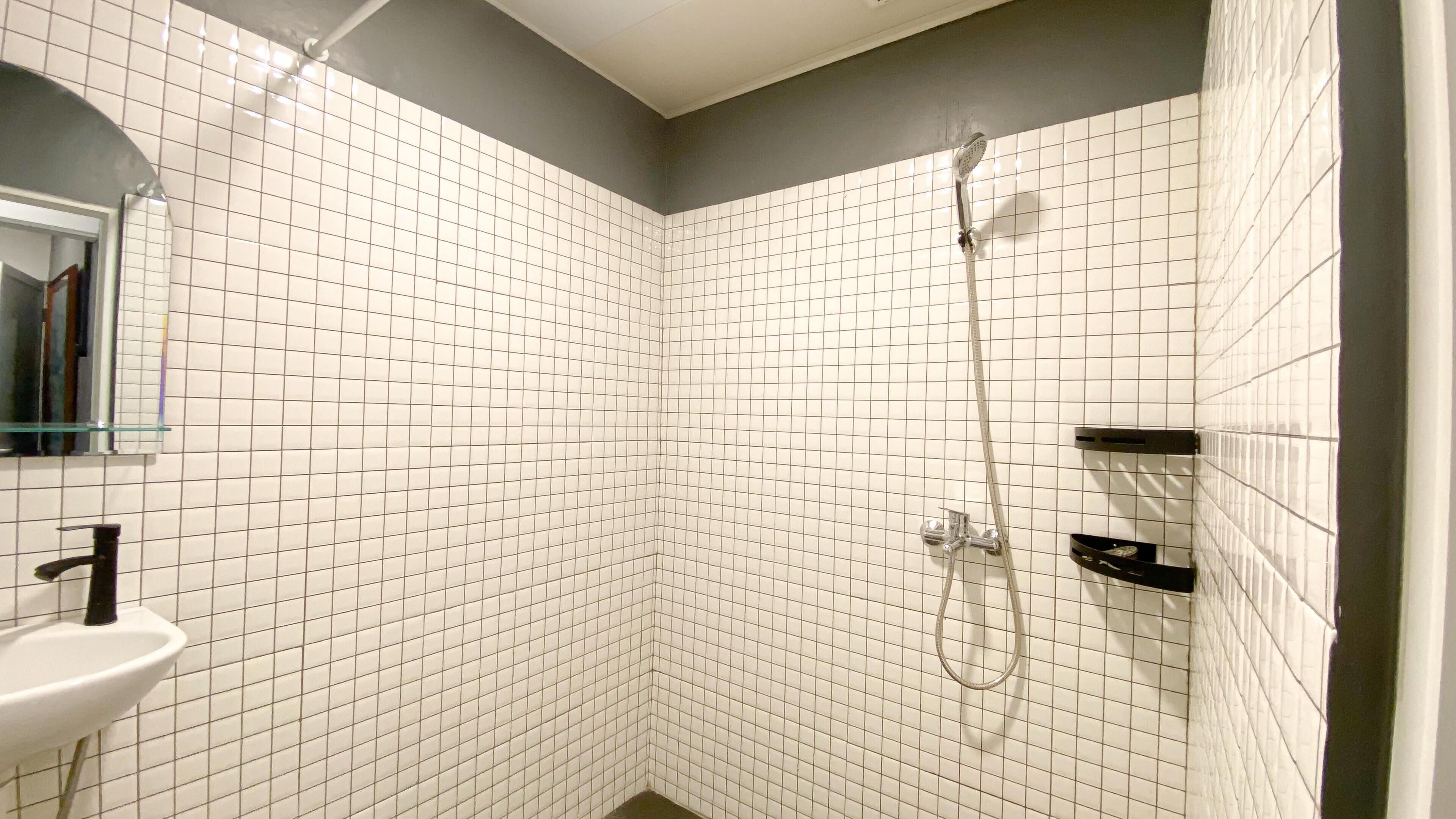 Ein Badezimmer im Eckbereich mit weißen Fliesen, einem Glasregal, einem Waschbecken und einer Dusche mit Duschbrause.