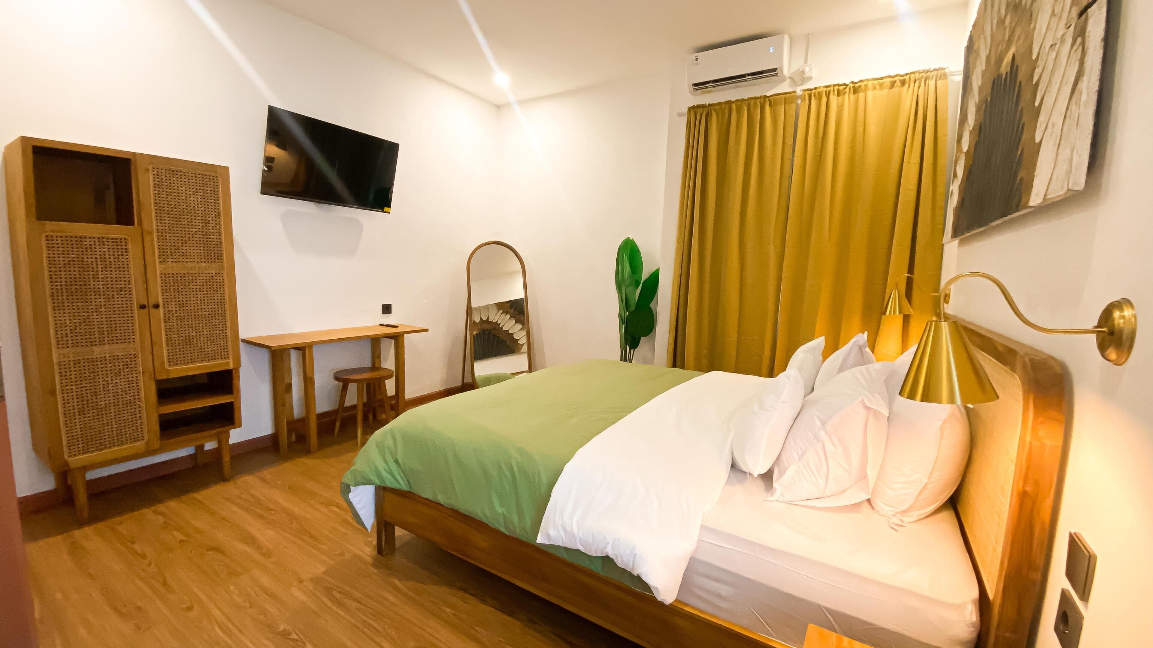 A hotel room with a bed, a desk, a mirror, a television, and an air conditioning unit.