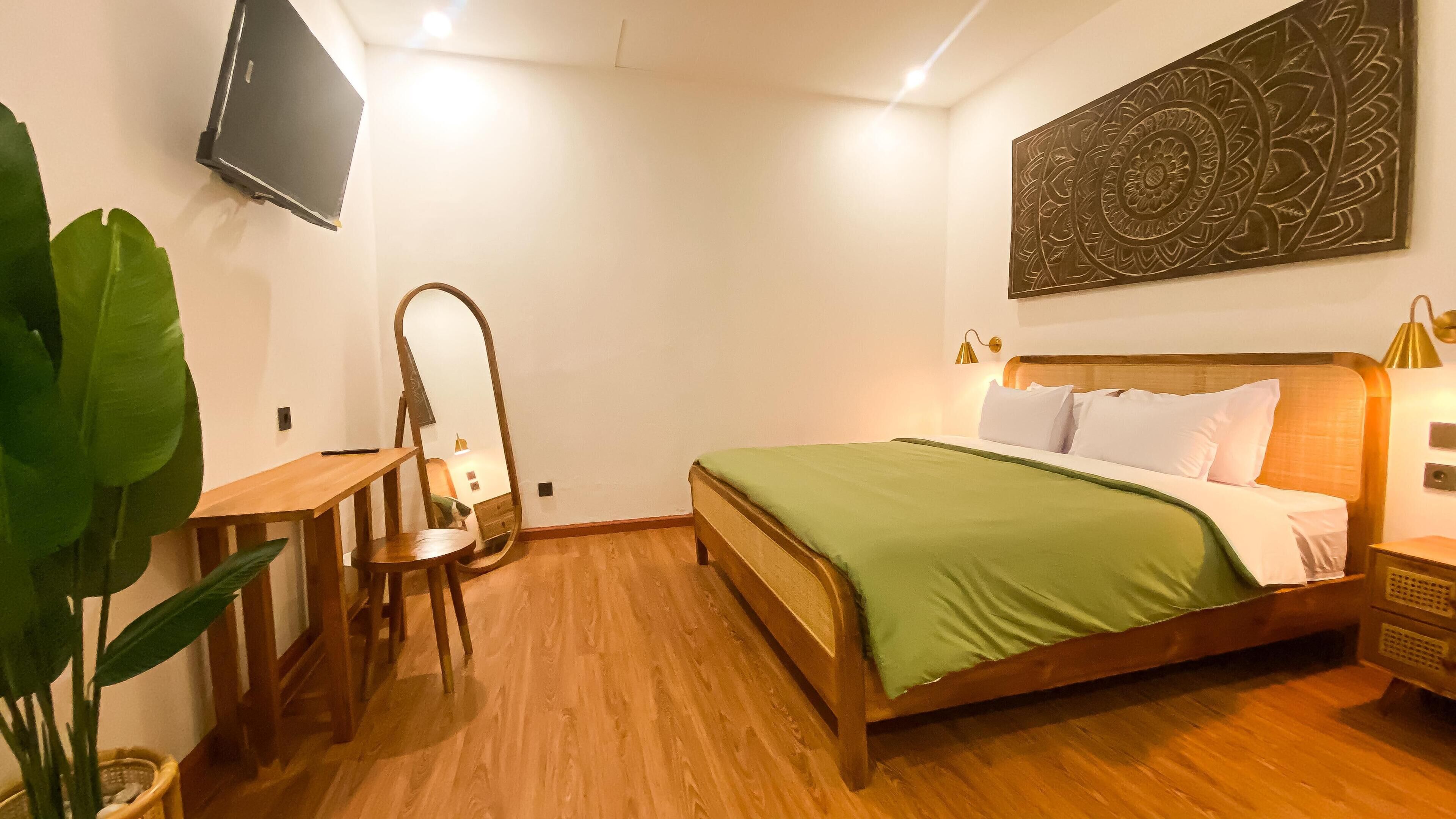 Ein Schlafzimmer mit einem hölzernen Bett, grüner Bettwäsche, einem Schreibtisch mit Stuhl, einem Spiegel, einem an der Wand befestigten Fernseher und einem Wandbild.