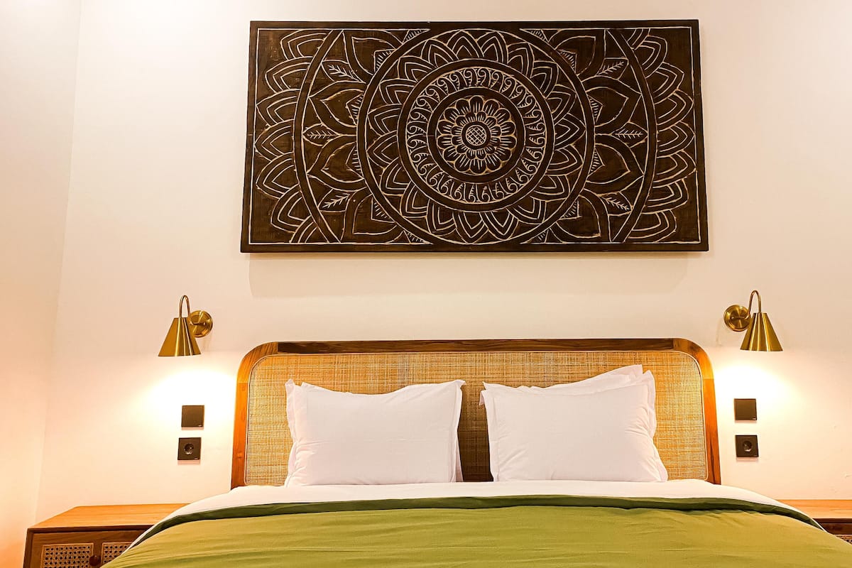 Un lit bien fait, recouvert d’une couvre-lit verte, avec deux oreillers blancs et une tête de lit à la texture tissée. Deux lampes murales sont fixées de part et d’autre du lit.