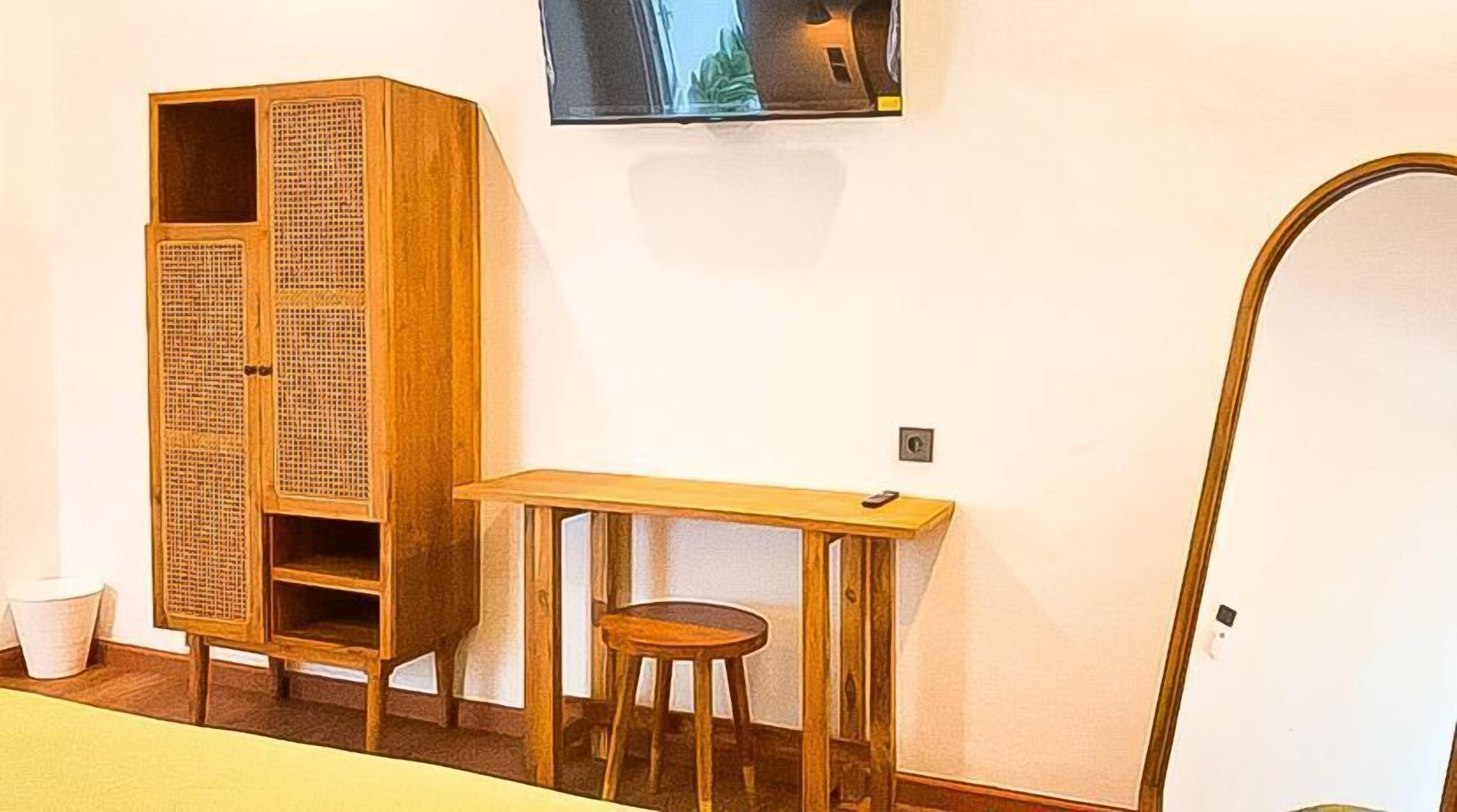 A room with a wooden wardrobe, a small table with a stool, a television mounted on the wall, and a door.