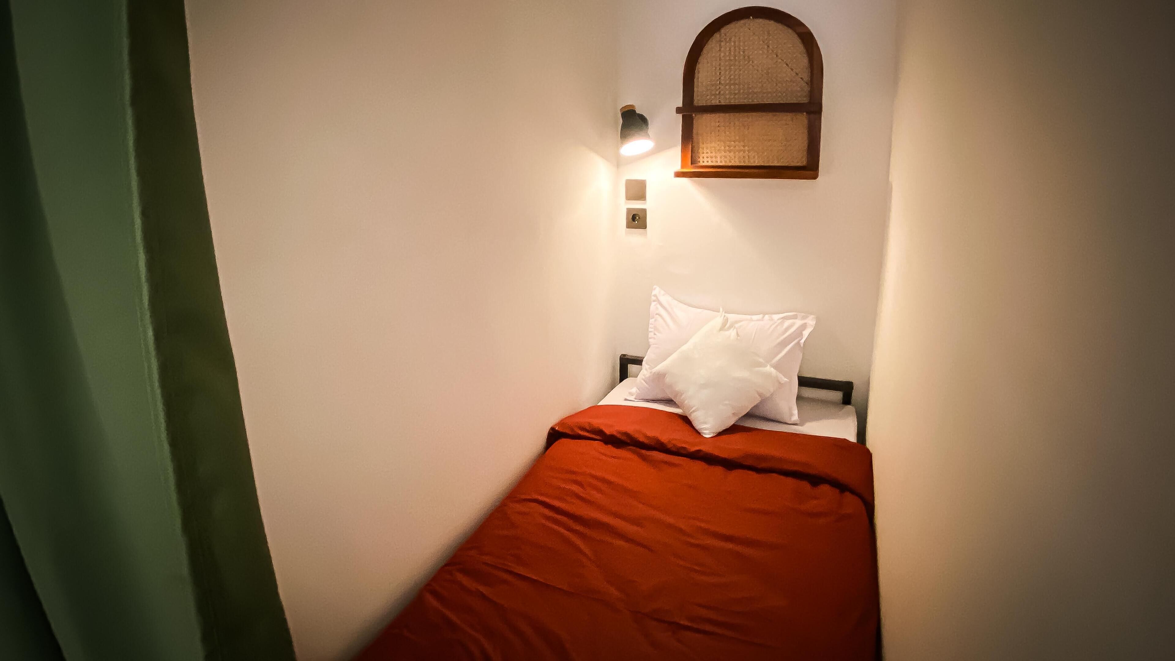 A small, single-bed room with a red bedspread, white pillows, a wall-mounted light, and a small window with a woven blind.
