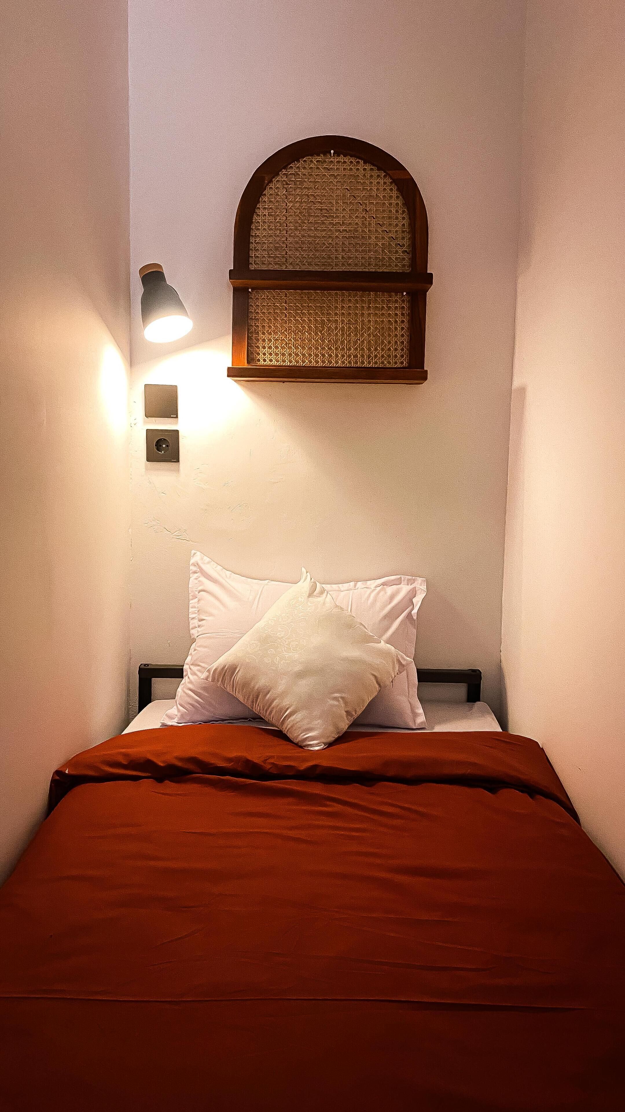 A single bed with a wooden headboard, a woven headboard above it, and a wall-mounted light fixture.