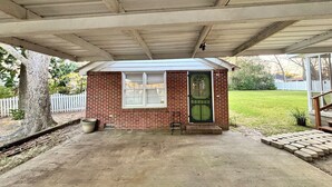 A small brick building with a green door and windows, under a covered area with a concrete floor.