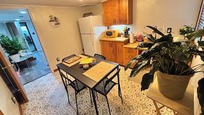 A compact kitchen with wooden cabinets, a refrigerator, and a microwave. There is a dining table with chairs, a potted plant, and a patterned floor.