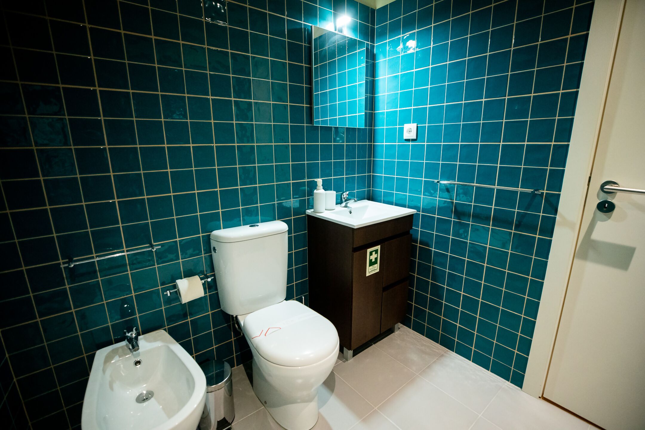 A bathroom with teal tiles, a white toilet, a bidet, and a sink with a cabinet.