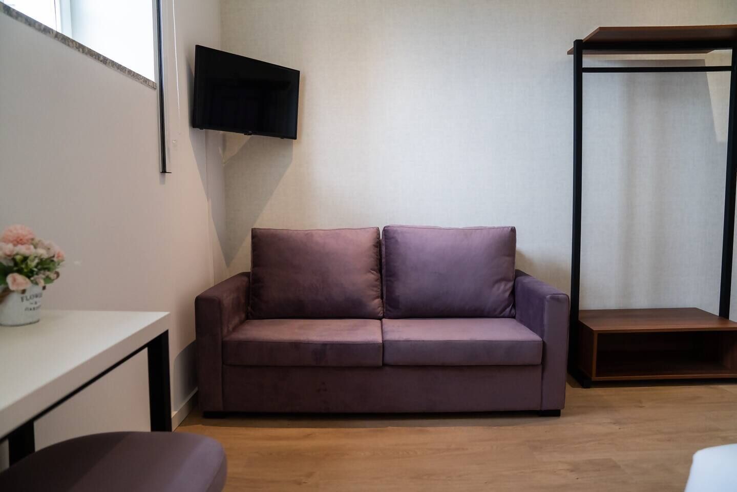 A room with a purple sofa, a small wooden table, a TV mounted on the wall, and a window with a flower vase.