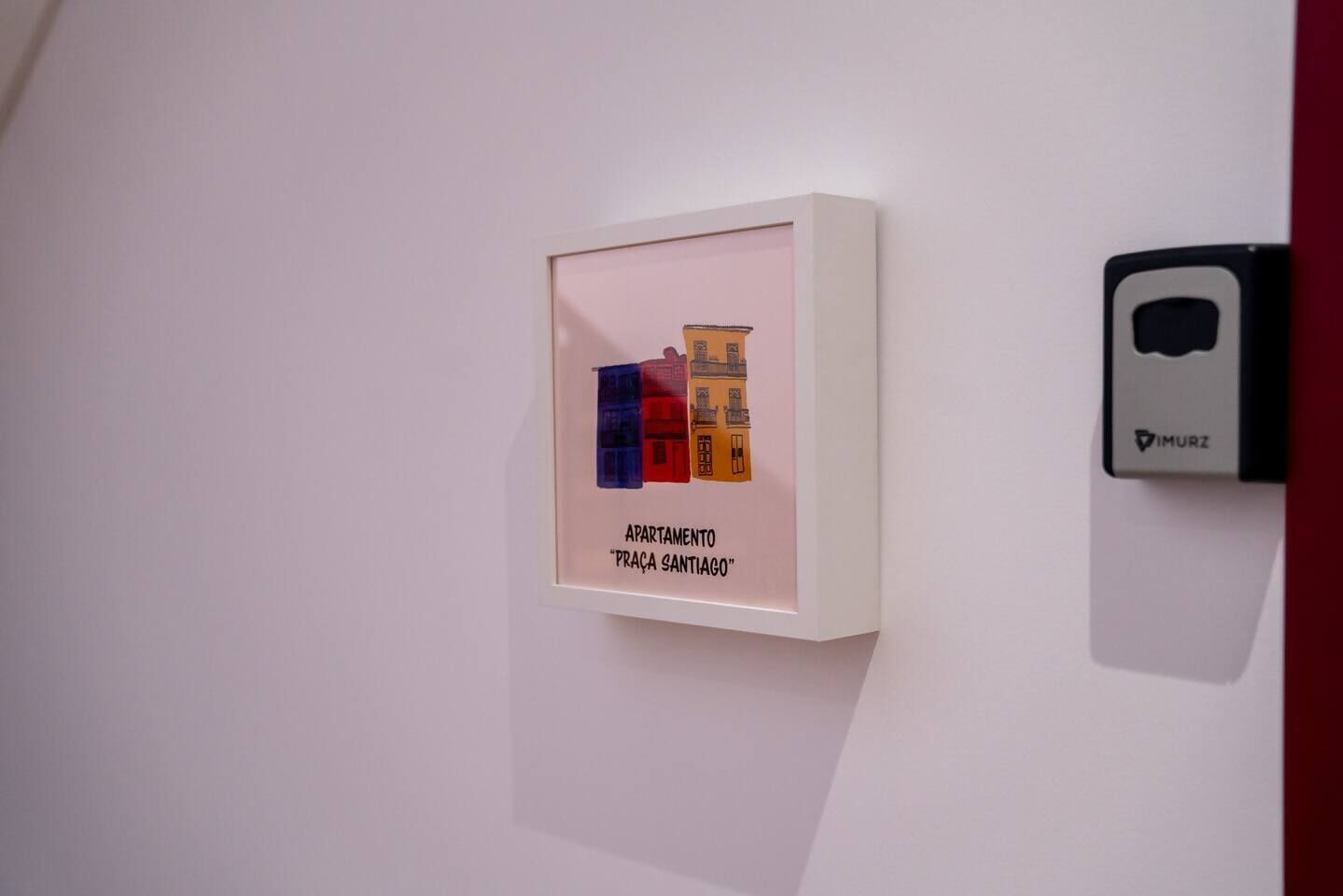 A framed artwork depicting colorful buildings with the text "APARTAMENTO PRAÇA SANTIAGO" on a white wall.