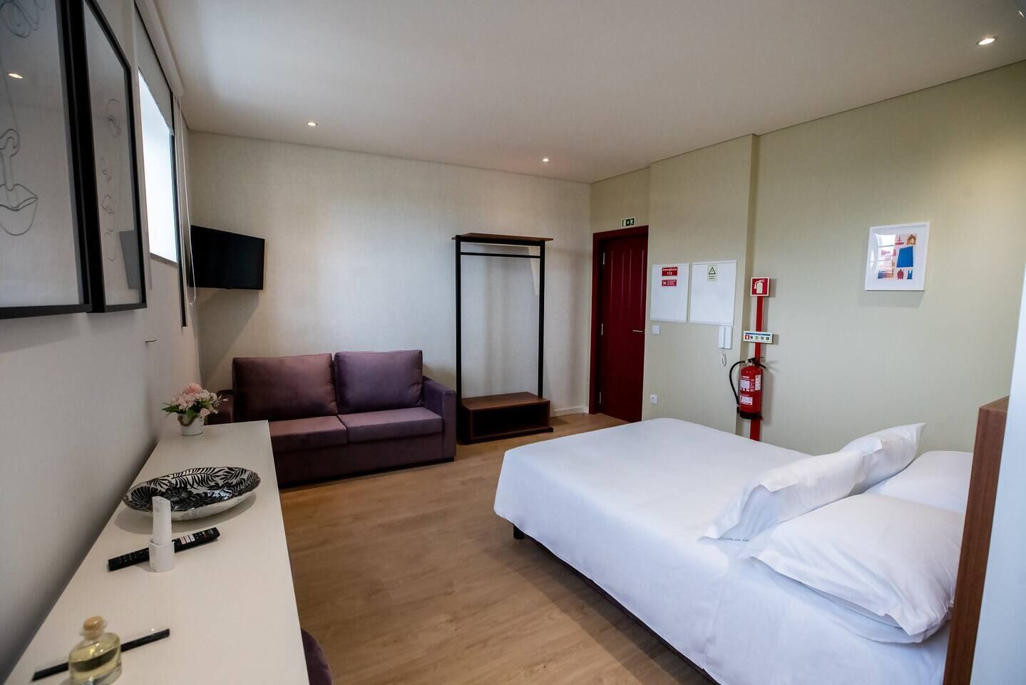 A hotel room with a bed, a sofa, a desk with a vase of flowers, a fire extinguisher, and a door.