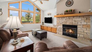 A living room with a stone fireplace, a ceiling fan, a large window, and a TV mounted on the wall. - Hot Tub, King Beds, Mountain Views on Balcony & Patio (Estes Park)
