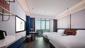 Une chambre d’hôtel comprenant un lit, un bureau, une chaise, une petite table avec un vase et une grande fenêtre donnant sur la ville.