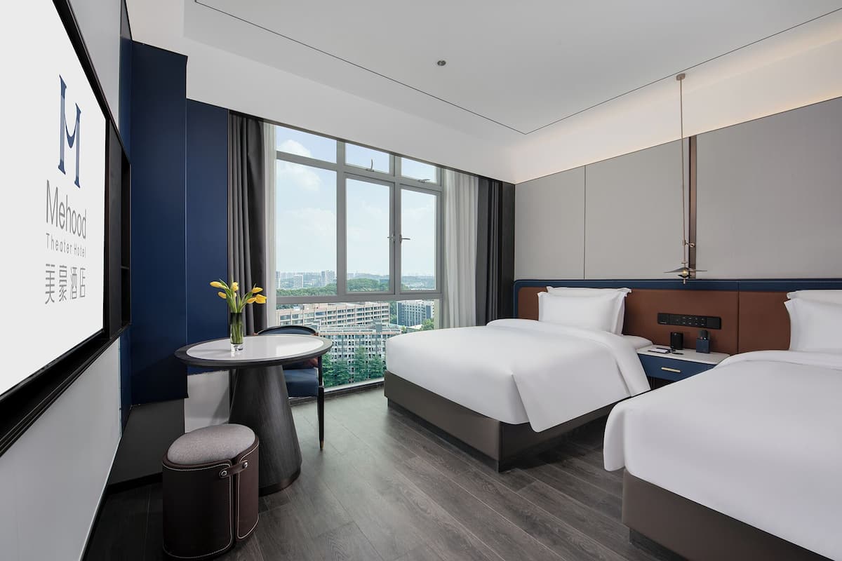 Habitación de hotel con dos camas, una mesita redonda con un florero y un ventanal con vistas a la ciudad.