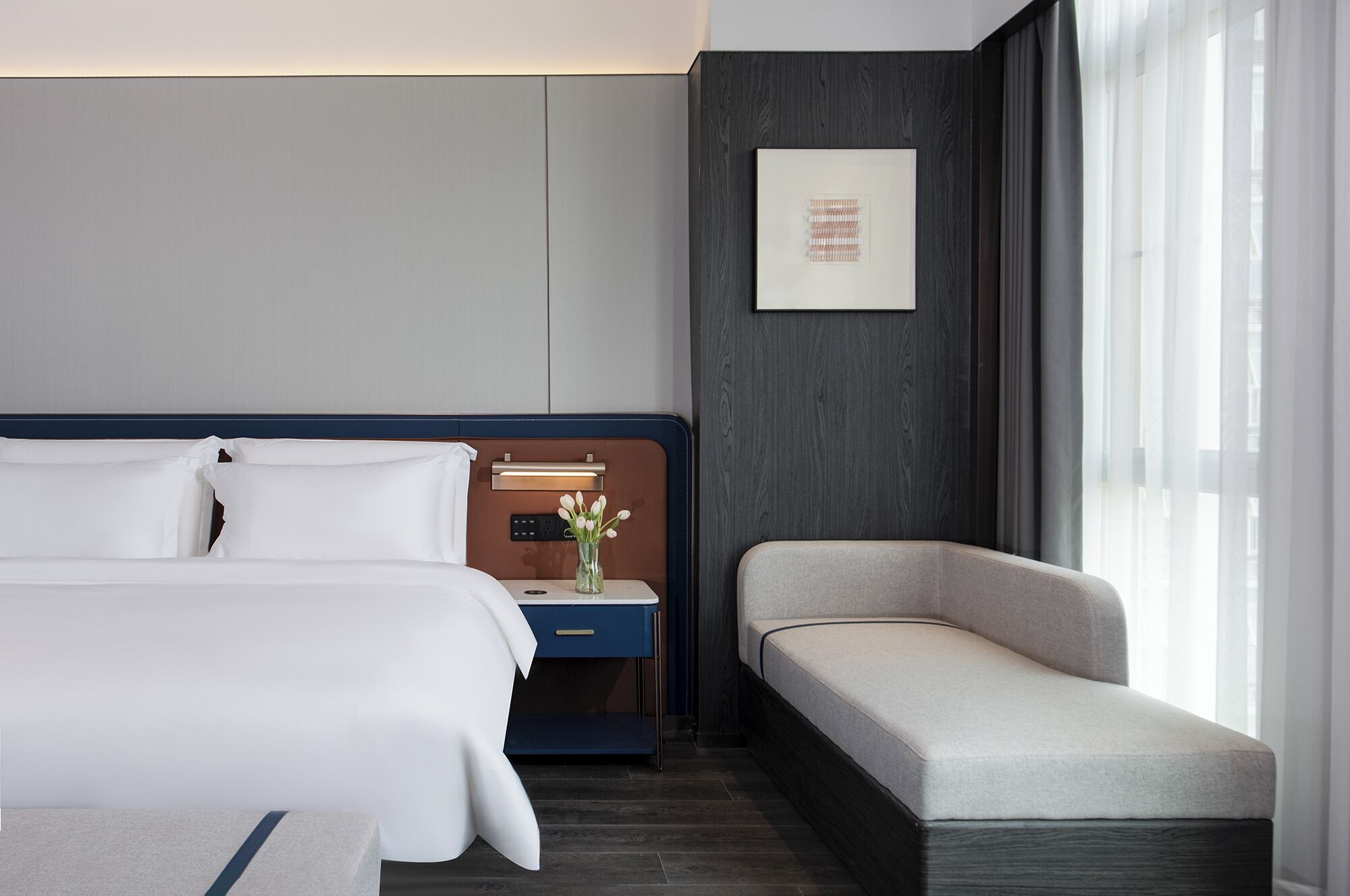 Ein Hotelzimmer mit einem großen Bett, einem Nachttisch mit Lampe, einem kleinen Tisch mit Blumenvase, einem Sofa und einem gerahmten Bild an der Wand.