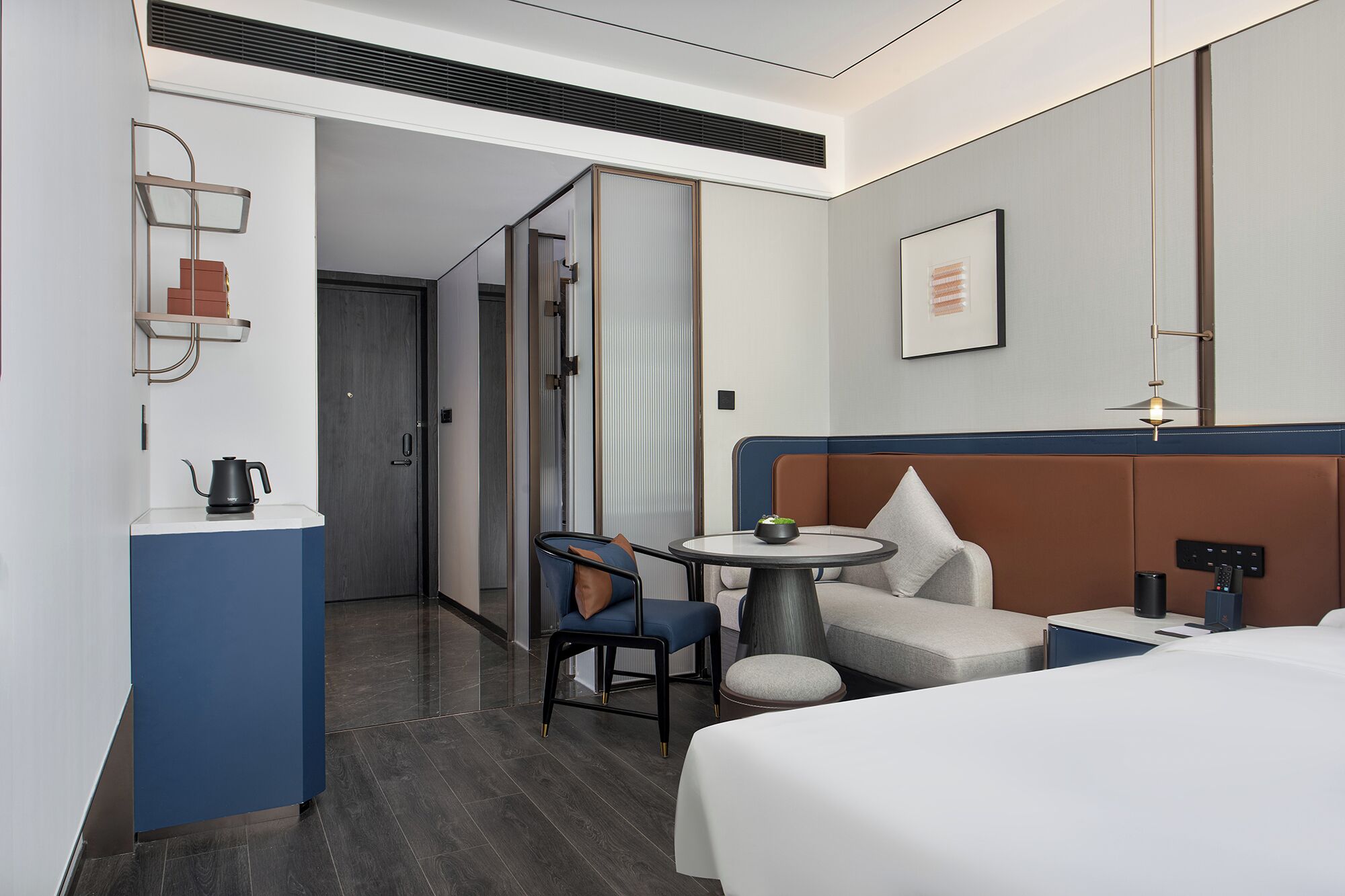 A modern hotel room with a bed, a small table with a bowl, a chair, a desk with a lamp, and a wardrobe.