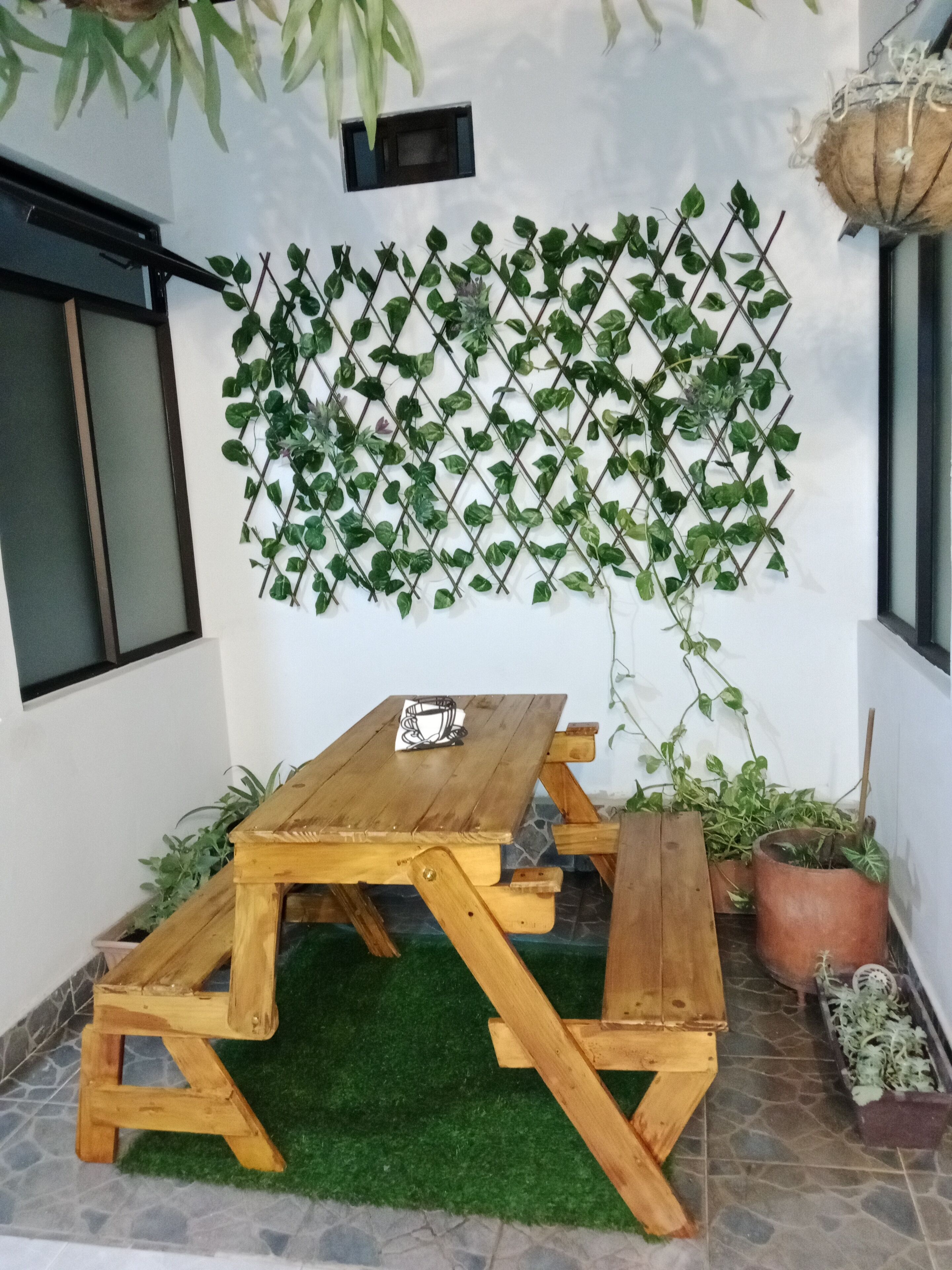 Une table de pique-nique en bois avec des bancs, un sol recouvert d’un gazon synthétique vert et une plante grimpante fixée au mur.