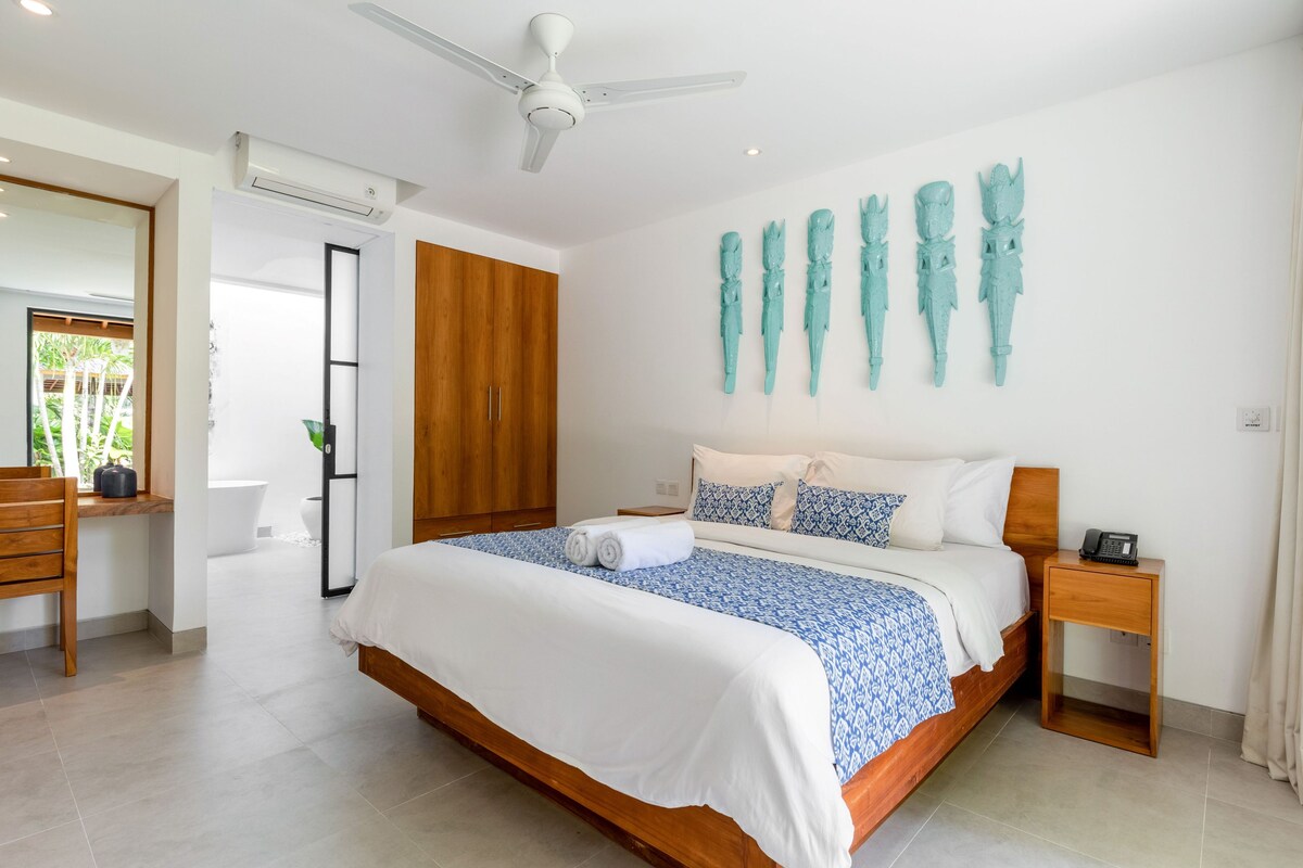 A modern bedroom with a large bed, wooden headboard, and a ceiling fan. There is a wooden nightstand with a phone and a wall-mounted sculpture.