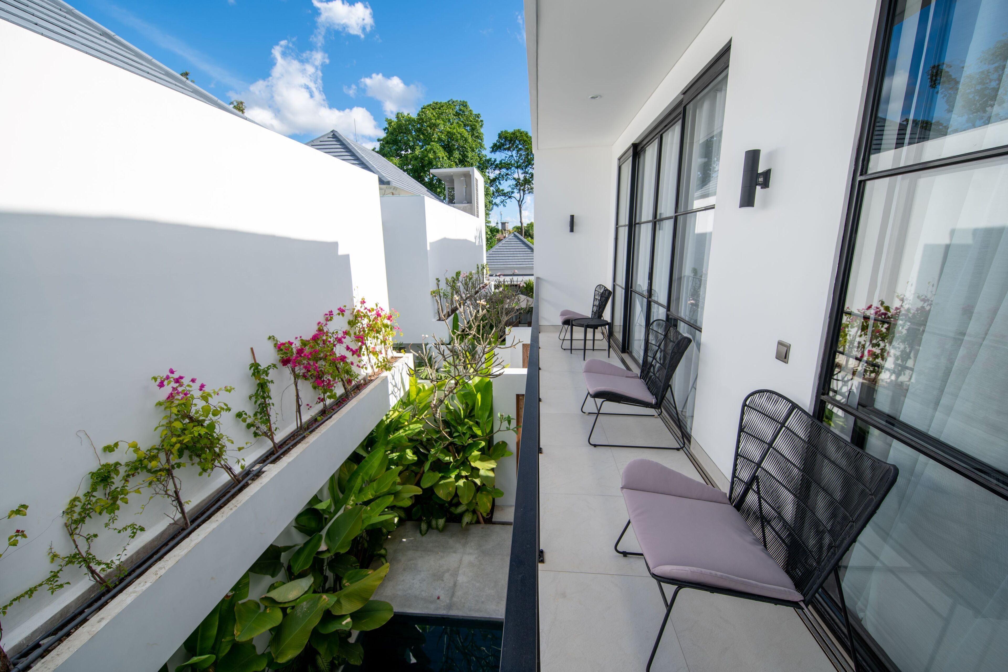 A narrow outdoor corridor with a white wall on one side and a glass door on the other. There are potted plants and flowers along the wall, and a few chairs are placed near the glass door.