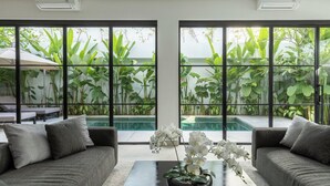 Un soggiorno moderno con divano, tavolino da caffè e un'elegante composizione floreale, e ampie finestre che si affacciano su una piscina e una vegetazione rigogliosa.