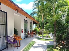 A modern building with a covered outdoor area featuring wooden tables and chairs, surrounded by lush greenery and palm trees.
