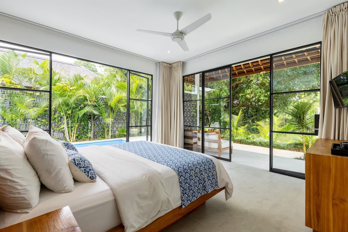 Ein Schlafzimmer mit Bett, Deckenventilator, großen Fenstern und Blick auf Grünflächen.