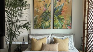 Une chambre à coucher avec un lit, des oreillers, une grande plante en pot et deux tableaux accrochés au mur.