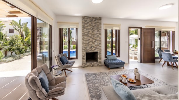 A spacious living area with a fireplace, comfortable seating, and large windows offering views of a pool and garden. - Ayia Napa Seaside Villa Ionion H15 (Sotira)